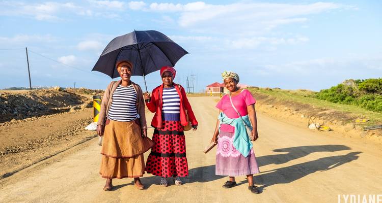 Drie lokale vrouwen staan glimlachend op een landelijke zandweg, een van hen houdt een paraplu vast onder felle zonneschijn.