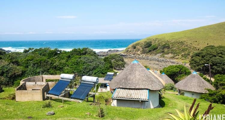 Rieten hutten kijken uit over een rotsachtige kustlijn en turquoise zee op een weelderige groene heuvelflank.