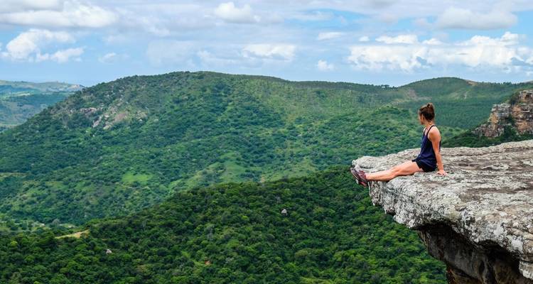 Een vrouw zit op de rand van een rotsachtige klif en kijkt uit over glooiende groene bergen en valleien.