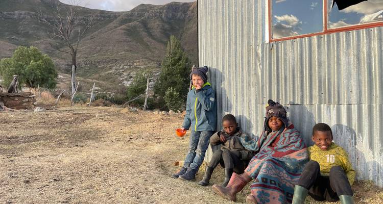 Vier kinderen die zitten en staan naast een golfplaten hut met berglandschap op de achtergrond.