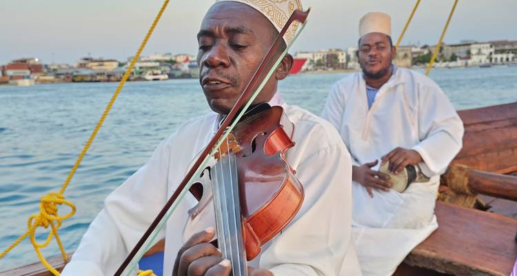 Muzikant die viool speelt op een traditionele houten boot met een andere man die begeleidt, kustplaats wazig op de achtergrond.