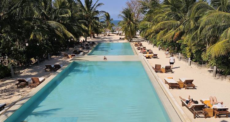 Luxe strandresort met twee lange overloopzwembaden in de schaduw van palmbomen en loungerende gasten.