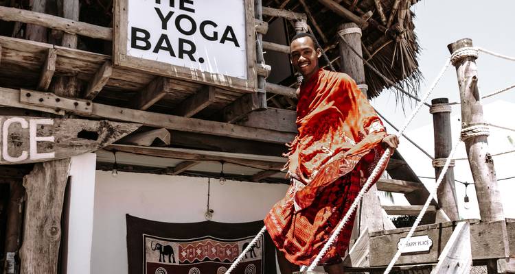 Glimlachende Maasai man in felgekleurde traditionele kleding staand op houten treden van een strandbar.