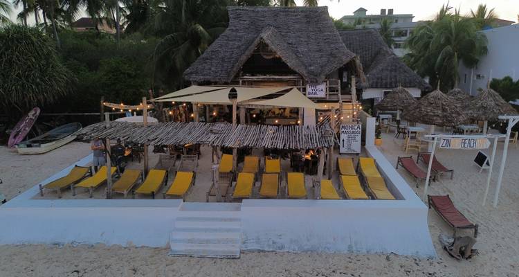 Luchtfoto van een strandrestaurant met gele ligstoelen en rieten dak omringd door palmbomen.