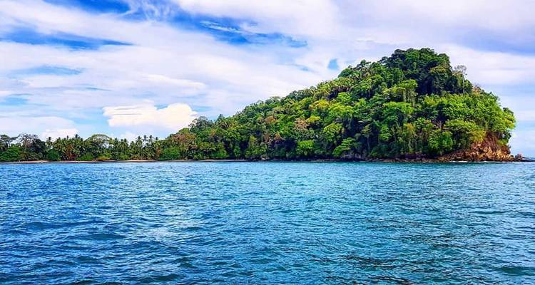 Exuberante isla tropical cubierta de árboles rodeada por un océano azul cristalino bajo un cielo parcialmente nublado.