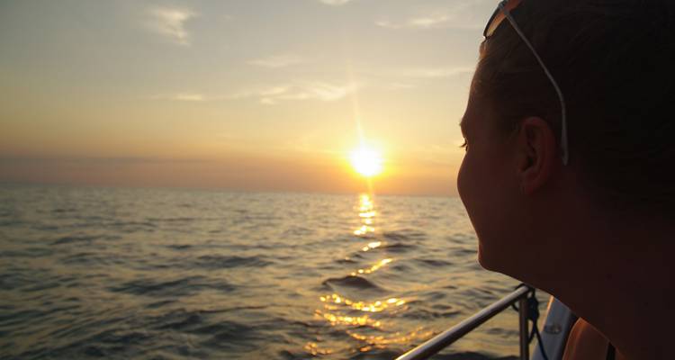 Persona en un bote mirando fijamente una puesta de sol naranja sobre un mar tranquilo, con pequeñas olas reflejando la luz.