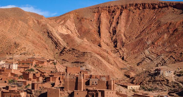 Village en terre niché sous des falaises rocheuses rouges abruptes dans une vallée de montagne aride.