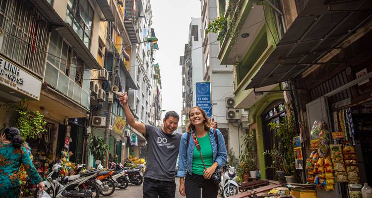 Lächelnde Reisende gehen durch eine enge Hanoi-Gasse, die mit Motorrollern, Geschaeften und hohen Wohnhaeusern gesaeumt ist.