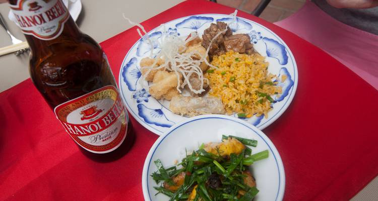 Repas vietnamien comprenant du riz frit, du porc, des en-cas croustillants et une bouteille de bière de Hanoï sur une nappe rouge.