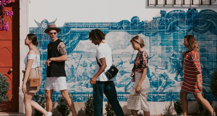 Young travellers stroll past a large blue azulejo tile mural on a sunny Portuguese street.
