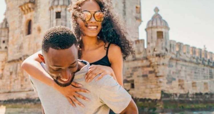 Mujer sonriente cargando a su pareja en la espalda frente a la Torre de Belém de piedra de Lisboa en un día despejado.
