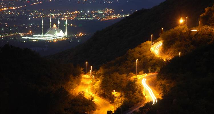 Vue nocturne en longue exposition d'une route sinueuse à flanc de colline avec des traînées lumineuses et la mosquée Faisal illuminée au loin en contrebas