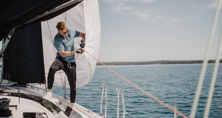 Un hombre ajustando una vela en la cubierta de un yate que se desliza por un mar azul cristalino.