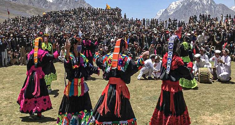 Bailarines tradicionales kalash actúan ante una gran multitud festiva con picos nevados al fondo.