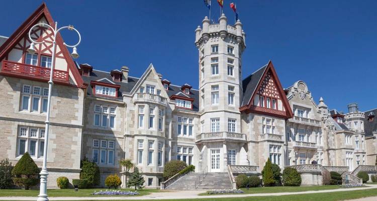 Groots stenen paleis met decoratieve torentjes en vlaggen onder een heldere blauwe hemel, omringd door verzorgde gazons