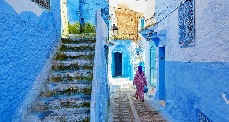 Smalle blauwe steegje met stenen trappen in Chefchaouen waar een vrouw in een gemusterde jurk wegloopt.