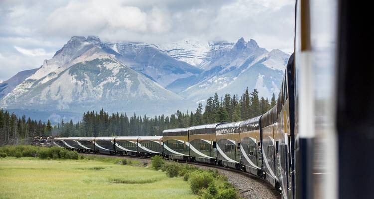 Un largo tren de pasajeros serpentea a través de un exuberante valle con las nevadas Montañas Rocallosas elevándose majestuosamente detrás.