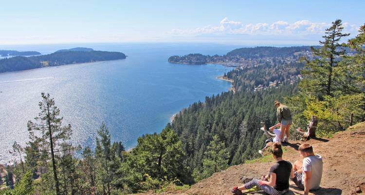 Grupo de senderistas descansando en un acantilado con vistas a una bahía brillante salpicada de penínsulas.