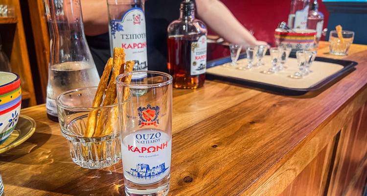 Mostrador de degustación con botellas de ouzo y tsipouro, vasos de chupito, palitos de pan y un anfitrión sirviendo licores.