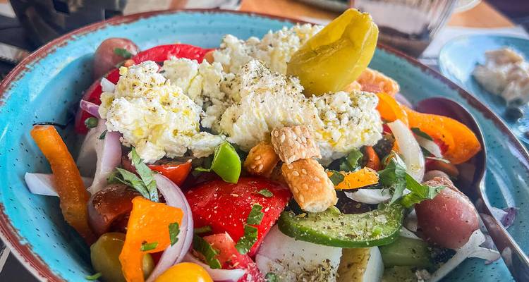 Ensalada griega vibrante en un tazón turquesa con queso feta, pimientos, pepinos, aceitunas, cebollas y palitos de pan.