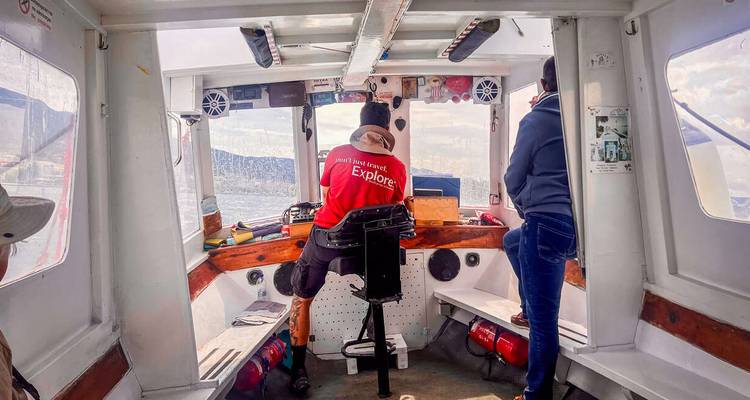 Interior de un pequeño barco de excursión; capitán vistiendo una chaqueta roja 'Explore' navega hacia mar abierto.
