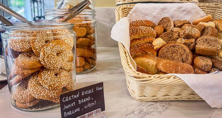 Frascos de vidrio y cesta de mimbre llenos de roscos de cebada cretenses tradicionales exhibidos sobre un mostrador de mármol.