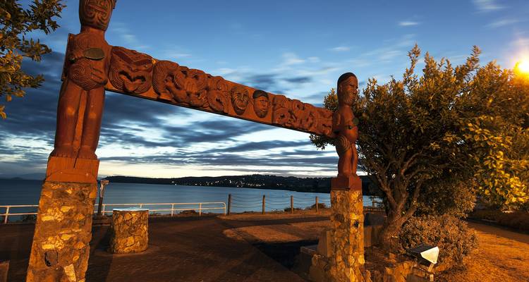 Sculptures maories sur un portail avec un lac et des collines en arrière-plan au crépuscule.