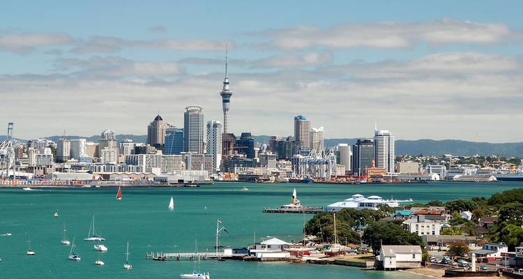 Klare Tagesansicht der Skyline von Auckland mit dem Sky Tower, der sich über den Hafen erhebt, und Segelbooten in türkisblauem Wasser.