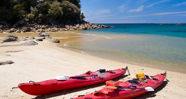 Rode zee-kajaks liggen op een stil zandstrand naast kristalhelder water en een rotsachtig schiereiland in Abel Tasman.