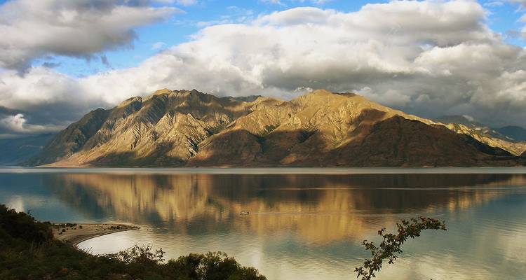 Cordillera bañada en luz dorada reflejada en un lago tranquilo bajo nubes dramáticas.
