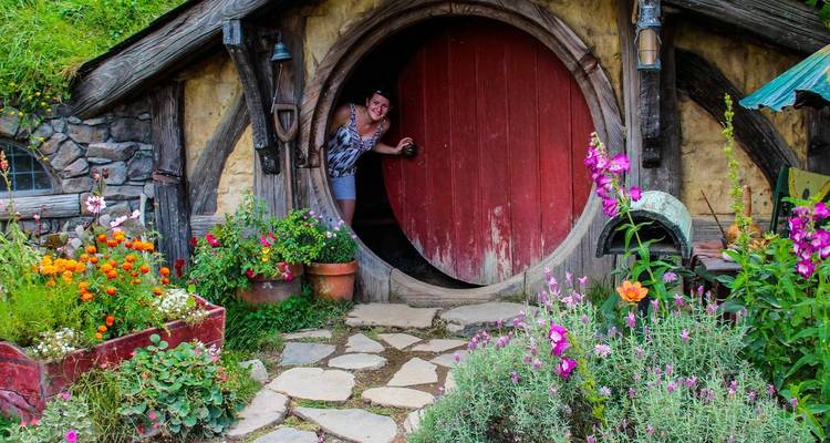 Una visitante asoma por una puerta roja redonda de Hobbit rodeada de flores coloridas y construcción de piedra rústica.