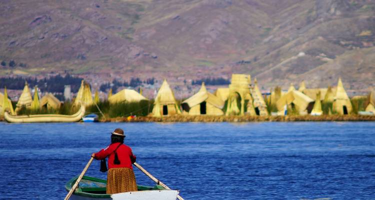 Woman in traditional dress rowing a small boat past reed island homes on Lake Titicaca.