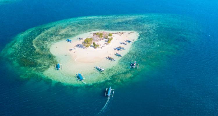 Luftaufnahme einer kleinen sandigen Insel, umgeben von türkisfarbenem Wasser mit Booten.