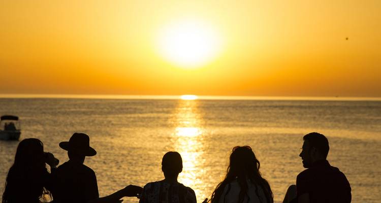 Des amis en silhouette regardent un coucher de soleil orange flamboyant sur les eaux calmes de la Méditerranée.