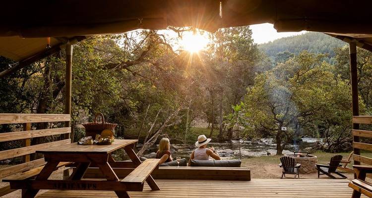 Relaxing camp setup with people by a river, surrounded by trees.
