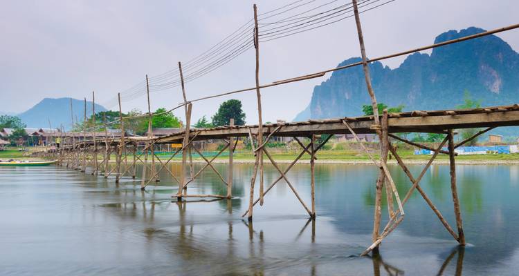 Estrecho puente peatonal de madera sobre pilotes atraviesa un río tranquilo con montañas kársticas de fondo