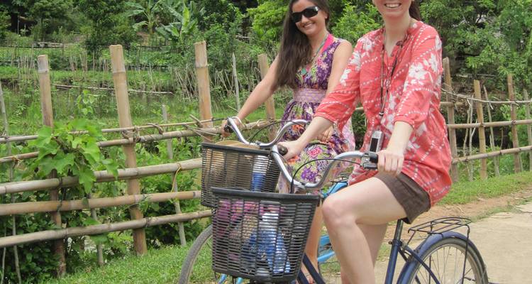 Dos viajeros sonrientes montando bicicletas por un sendero rural bordeado por cercas de bambú y vegetación.