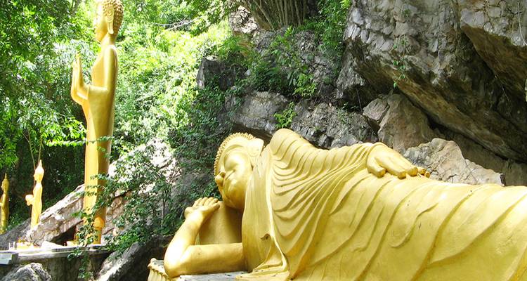 Estatua dorada de Buda reclinado anidada contra una ladera rocosa cubierta de árboles con figuras adicionales de Buda de pie cerca.