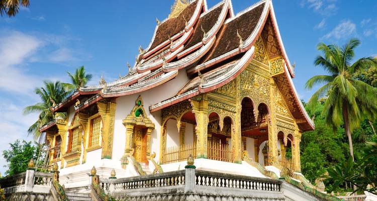Üppig geschmückter buddhistischer Tempel mit geschwungenem mehrstöckigem Dach und goldenen Schnitzereien, eingebettet zwischen Palmen unter einem strahlenden Himmel.