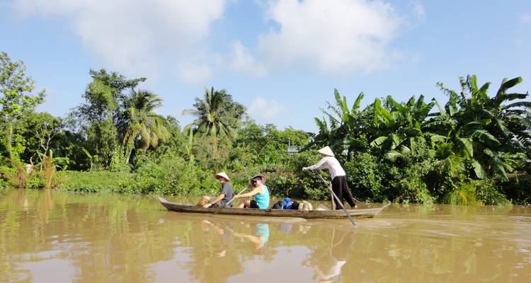 Holzboot gleitet entlang eines schlammigen tropischen Flusses vorbei an dichten Bananenbäumen mit drei Reisenden in konischen Hüten, die die Fahrt genießen.