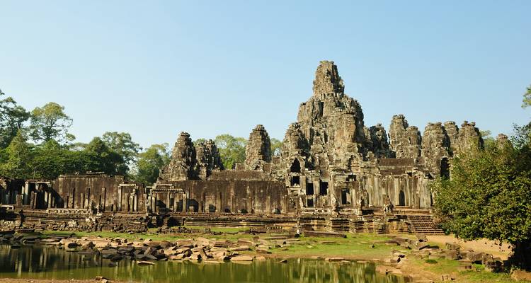 Anciennes tours de pierre et ruines du temple Bayon reflétées dans un petit étang sous un ciel clair.