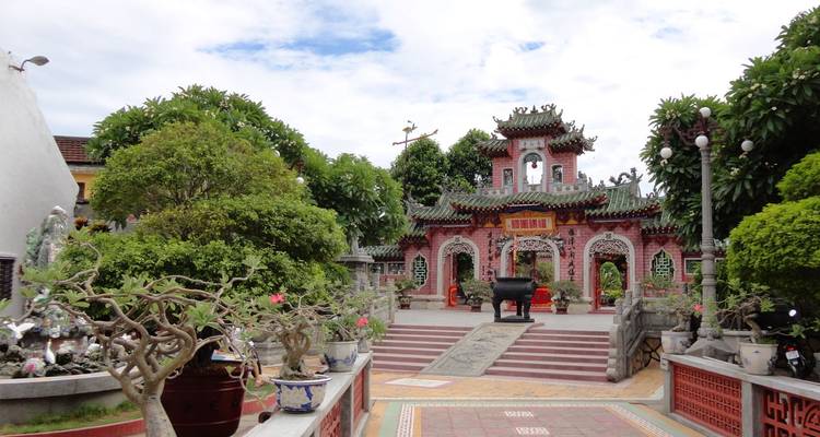 L'entrée ornée rose et verte d'une salle d'assemblée chinoise à Hoi An est encadrée par des bonsaïs et des pots en céramique sous un ciel nuageux parsemé.