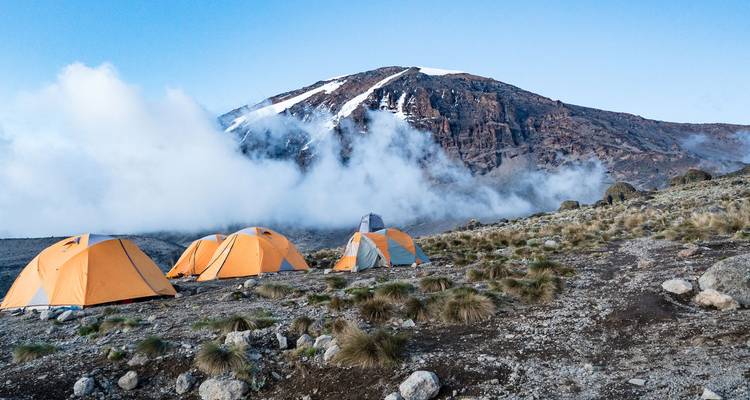 Orangefarbene Zelte auf einem Campingplatz mit dem Kilimandscharo, der teilweise in Wolken gehüllt ist.