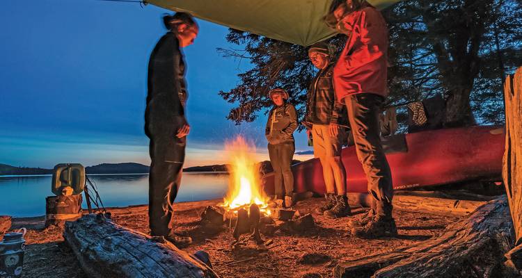 Grupo de personas alrededor de una fogata junto a un lago al atardecer.