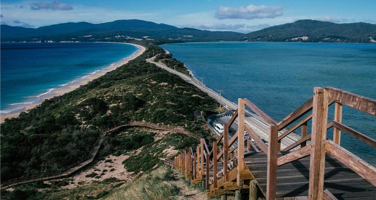 Escalera de madera bajando hacia un largo istmo arenoso que divide las aguas azul profundo del océano con colinas boscosas ondulantes a lo lejos.