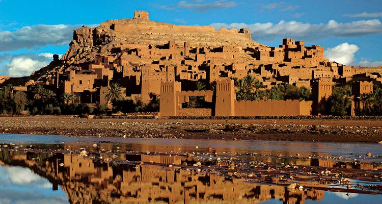 Het aarden citadel van Ait Benhaddou rijst boven een rivierbedding uit, weerspiegeld in het stille water onder een felblauwe hemel.