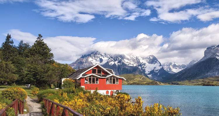 A red lakeside lodge framed by golden shrubs with dramatic snowy peaks across turquoise waters in Patagonia.