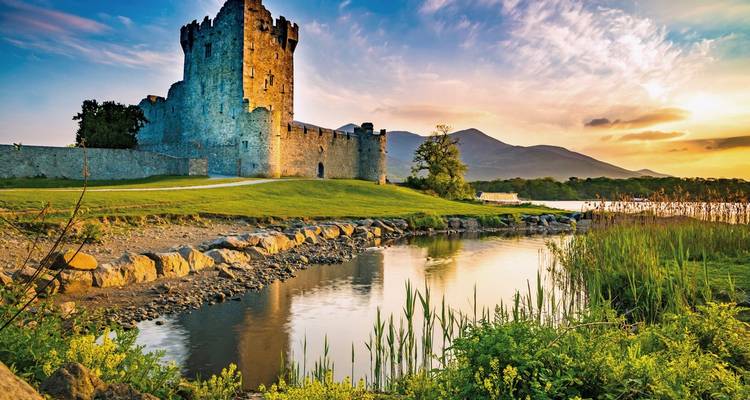 Picturesque Ross Castle beside a tranquil stream at sunset with mountains in the distance.