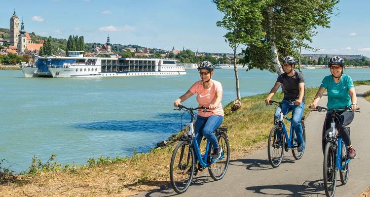 Radfahrer fahren an einem Fluss entlang mit einem Kreuzfahrtschiff und einer historischen Stadt in Sicht.