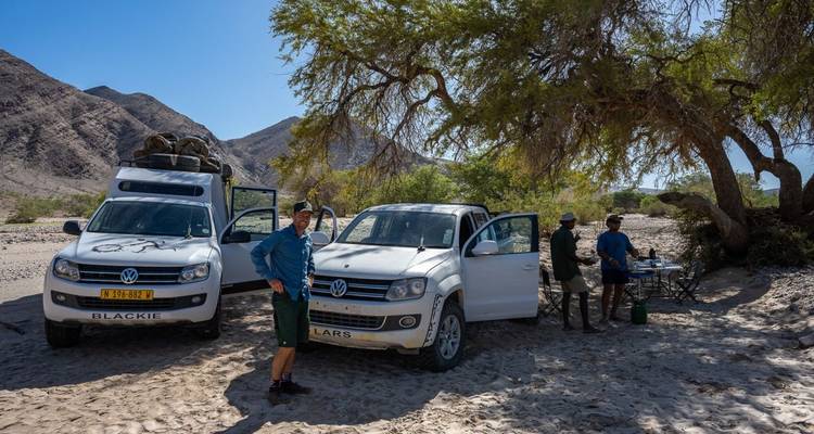 Grupo de personas paradas cerca de automóviles con arena y montañas al fondo.